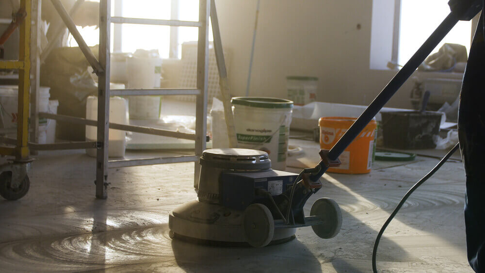 a worker using a concrete polishing machine to polish the floor of a house that has painting buckets on the floor