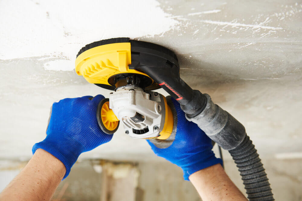 a worker concrete polishing the roof of a concrete room with a hand held grinder that has a dust extraction hose coming off of it