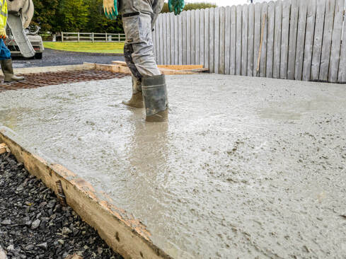 A concreting Ballarat worker standing in a bed of wet concrete that is forming a driveway with concrete boots on and a concrete truck in the background